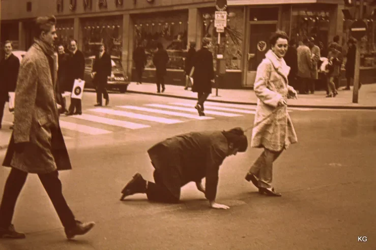 Vintage sepia street scene showing a man crawling on hands and knees across a crosswalk while two well-dressed women walk nearby.